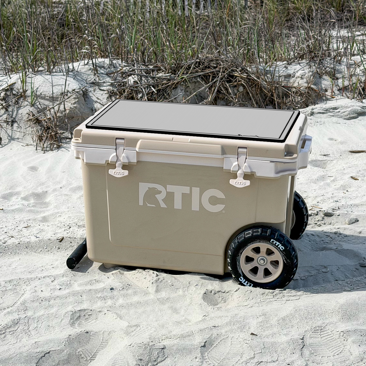 A beige RTIC cooler with a black slatted lid and black wheels, sitting on white sand in an outdoor beach setting with grass in the background with a DEKit foam grey over black marlin design on top.