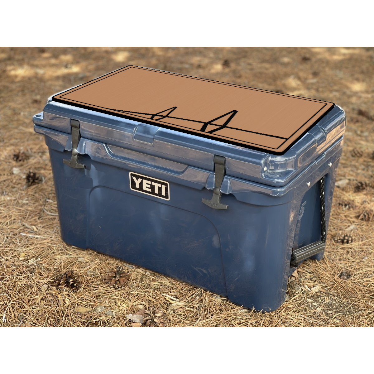 A blue YETI cooler with a DEKit cooler pad adhered to the lid, shown outdoors on a bed of dry pine needles and pinecones. The YETI logo is clearly visible on the front.