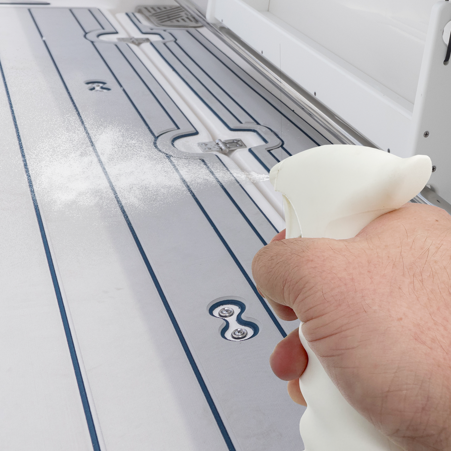 A hand holding DEKit's All-Purpose Cleaner spray bottle and spraying a fine mist onto a ice grey deck mat with navy blue teak lines.
